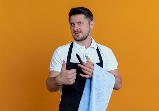 homem barbeiro de avental com a toalha na mão segurando o pincel de barbear com espuma e navalha sorrindo mostrando os polegares em pé sobre a parede laranja