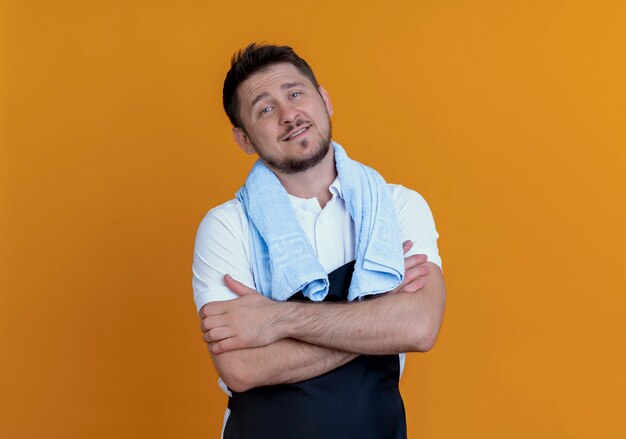 Homem barbeiro de avental com uma toalha em volta do pescoço lookign para a câmera sorrindo confiante com os braços cruzados em pé sobre um fundo laranja