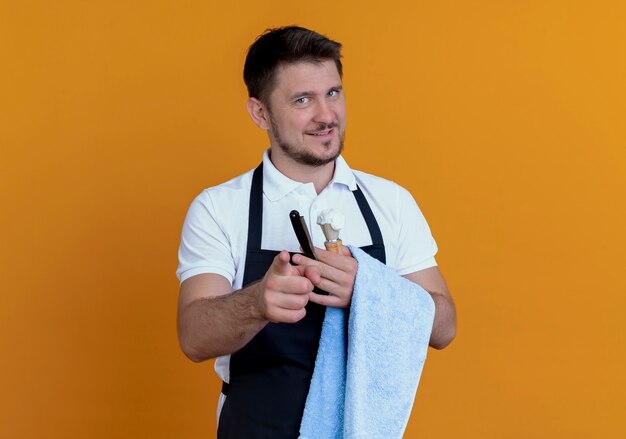 homem barbeiro de avental com uma toalha na mão segurando um pincel de barbear com espuma e navalha sorrindo confiante em pé sobre a parede laranja