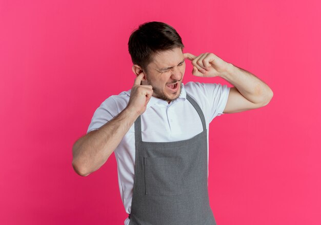 Homem barbeiro de avental fechando as orelhas com os dedos e uma expressão irritada em pé sobre um fundo rosa