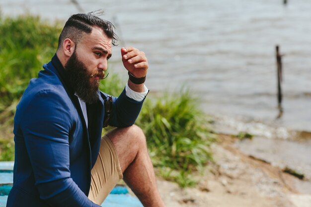 Homem barbudo americano parece na margem do rio com uma jaqueta azul