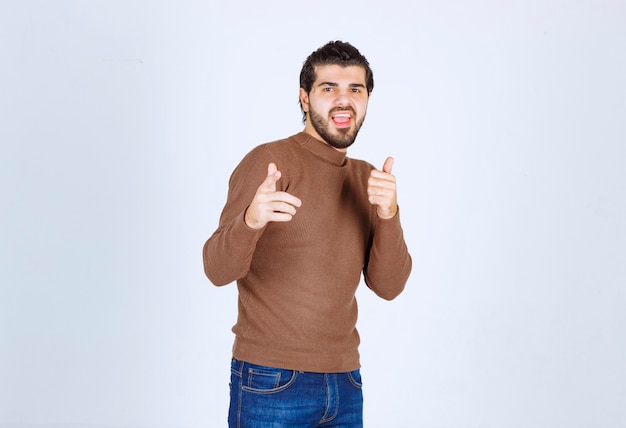 Homem barbudo animado de suéter marrom, apontando os dedos para a câmera isolada em um fundo branco. Foto de alta qualidade