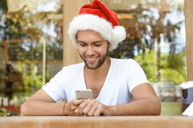 Homem barbudo atraente com chapéu de Papai Noel celebrando o Natal em um país tropical, verificando redes sociais e lendo mensagens