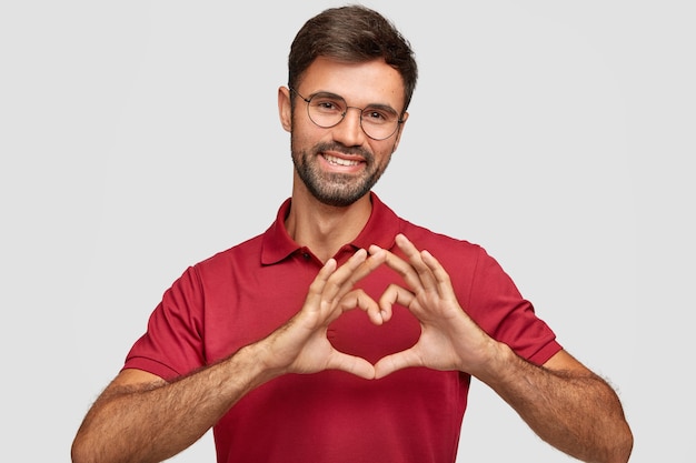 Foto grátis homem barbudo atraente e amigável faz gestos de coração, sorri agradavelmente, usa óculos e camiseta vermelha, expressa amor