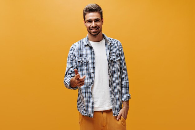 Homem barbudo bonito em calças laranja, camiseta branca e camisa quadriculada sorri sinceramente e aponta para a câmera em fundo laranja