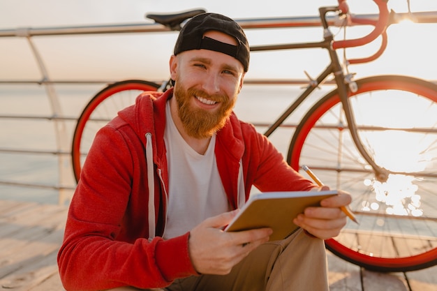 Homem barbudo bonito estilo hippie com capuz vermelho estudando freelancer online, escrevendo, fazendo anotações com uma bicicleta ao nascer do sol da manhã à beira-mar estilo de vida ativo e saudável