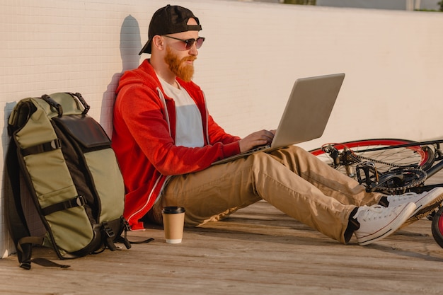 Foto grátis homem barbudo bonito estilo hippie trabalhando online freelancer no laptop com mochila e bicicleta no nascer do sol da manhã à beira-mar estilo de vida ativo saudável mochileiro viajante