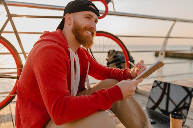 Homem barbudo bonito estilo hipster estudando freelancer online escrevendo fazendo anotações com mochila e bicicleta na manhã do nascer do sol à beira-mar mochileiro viajante estilo de vida ativo saudável