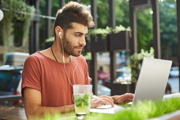 Homem barbudo bonito, freelancer trabalhando remoto em um café ao ar livre, programador com laptop ouvindo música para se concentrar no trabalho