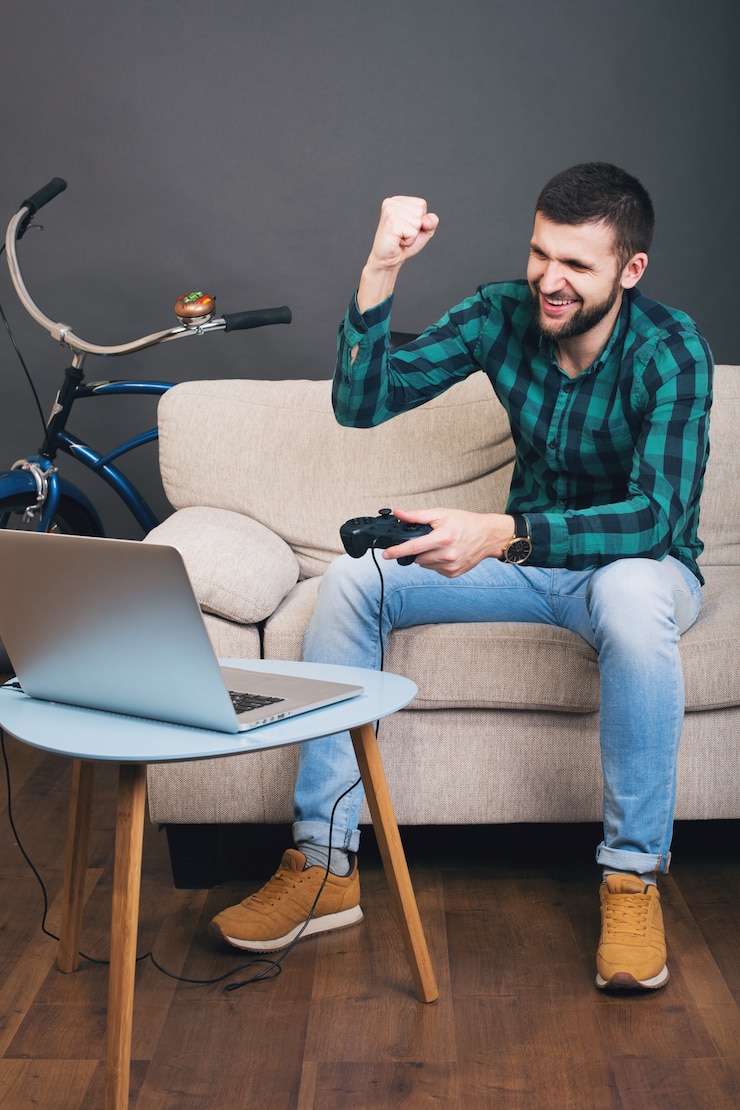 Foto grátis homem barbudo bonito jovem hippie sentado no sofá em casa, jogando videogame no notebook