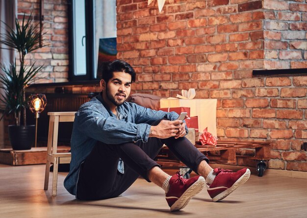 Homem barbudo casual indiano usando um smartphone em uma sala de loft com decoração de Natal.