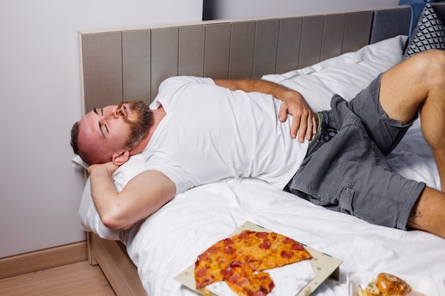 Homem barbudo caucasiano deita-se depois de comer pizza e hambúrgueres