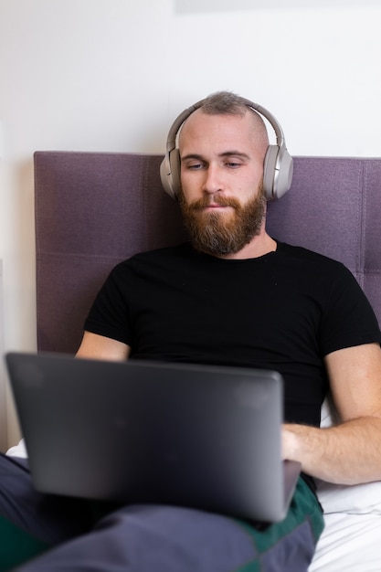 Homem barbudo caucasiano em fones de ouvido no quarto na cama, trabalhando no laptop em casa, digitando, pensando.