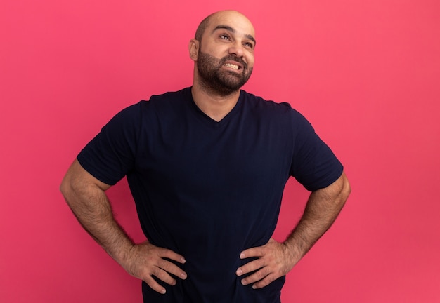 Foto grátis homem barbudo com camiseta azul marinho olhando para o lado com um sorriso no rosto e os braços na altura do quadril em pé sobre a parede rosa