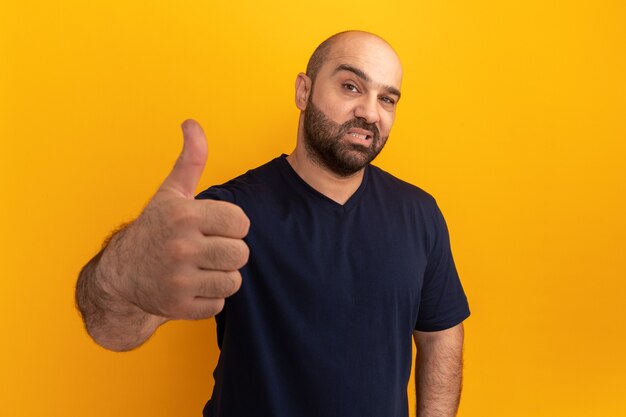 Homem barbudo com camiseta azul marinho sorrindo e mostrando os polegares em pé sobre uma parede laranja