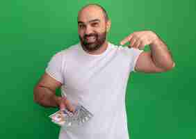 Foto grátis homem barbudo com camiseta branca segurando dinheiro feliz e positivo apontando com o dedo indicador para o dinheiro em pé sobre a parede verde