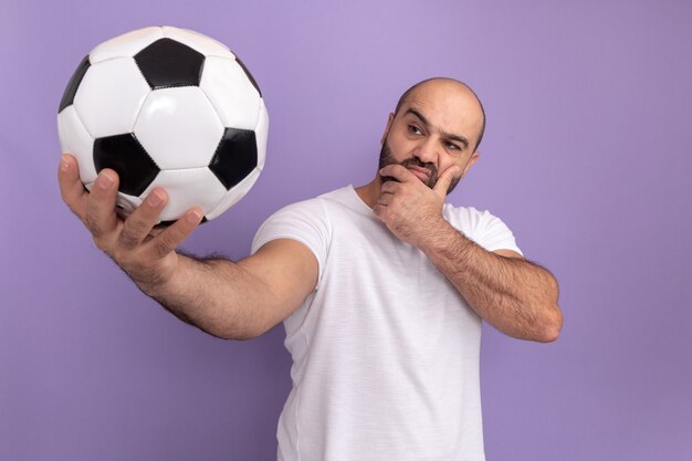 Homem barbudo com camiseta branca segurando uma bola de futebol olhando para ela com expressão pensativa no rosto pensando em pé sobre a parede roxa