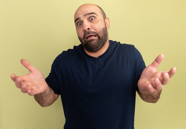 Homem barbudo com camiseta preta confuso e assustado com os braços estendidos em pé sobre a parede verde