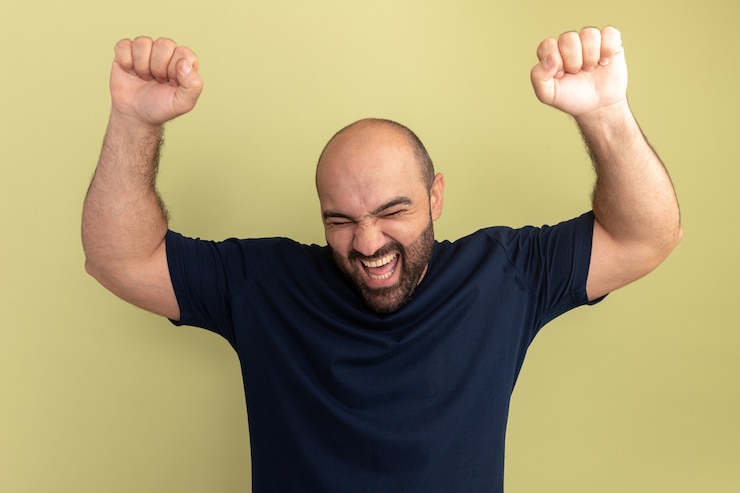 Foto grátis homem barbudo com camiseta preta louco, feliz e animado, gritando com os punhos cerrados em pé sobre a parede verde