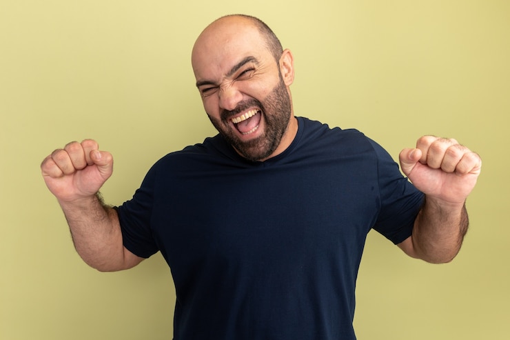 Foto grátis homem barbudo com camiseta preta louco, feliz e animado, gritando com os punhos cerrados em pé sobre a parede verde