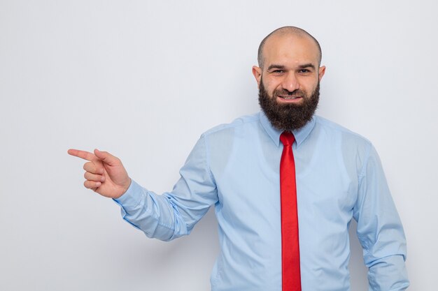 Homem barbudo com gravata vermelha e camisa azul, sorrindo alegremente e apontando com o dedo indicador para o lado