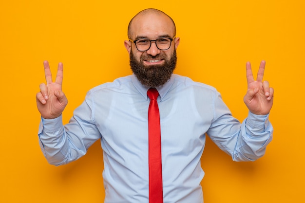 Homem barbudo com gravata vermelha e camisa azul usando óculos, olhando para a câmera, sorrindo alegremente mostrando o sinal V em pé sobre um fundo laranja