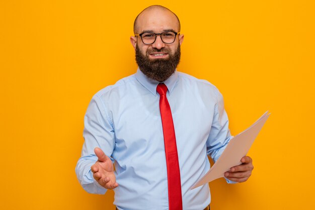 Homem barbudo com gravata vermelha e camisa azul usando óculos, segurando documentos, olhando oferecendo saudação de mão