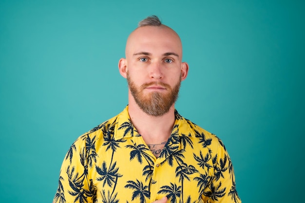Homem barbudo com uma camisa e uma estampa de palmeiras em uma parede turquesa olhando atentamente para a frente