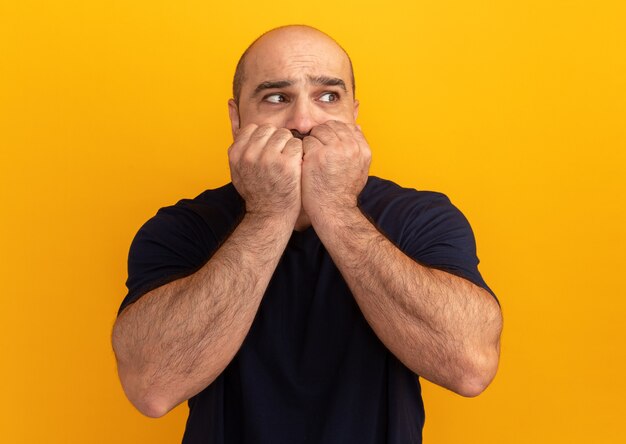 Homem barbudo com uma camiseta azul marinho olhando de lado estressado e nervoso roendo as unhas em pé sobre a parede laranja