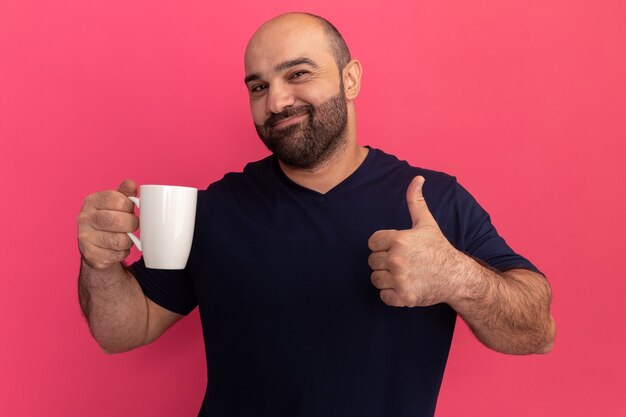 Homem barbudo com uma camiseta azul marinho segurando uma xícara, feliz e positivo, mostrando os polegares em pé sobre a parede rosa