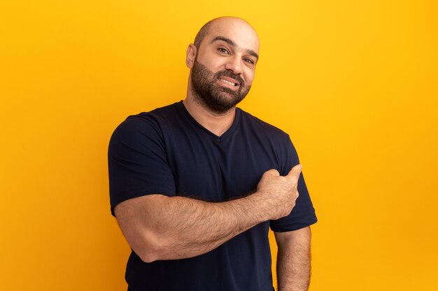 Homem barbudo com uma camiseta azul marinho sorrindo confiante apontando para trás em pé sobre a parede laranja