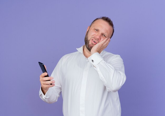 Homem barbudo de camisa branca segurando um smartphone parecendo confuso em pé sobre uma parede azul