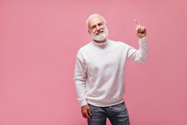 Homem barbudo de jeans e suéter apontando para cima na parede rosa