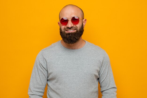 Homem barbudo de moletom cinza com óculos vermelhos, olhando para a câmera, feliz e positivo, sorrindo amplamente em pé sobre um fundo laranja