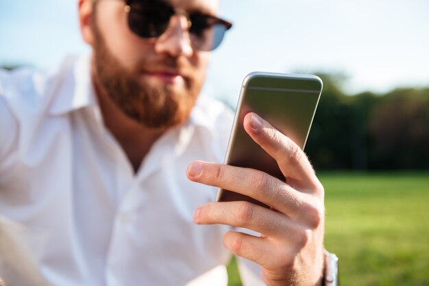 homem barbudo de óculos escuros e camisa enquanto estiver usando o smartphone. Concentre-se no telefone