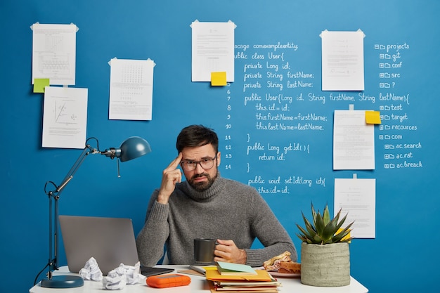 Homem barbudo de óculos pondera sobre o projeto de inicialização, tem expressão infeliz, tenta se concentrar, bebe café, faz trabalho remoto no próprio armário