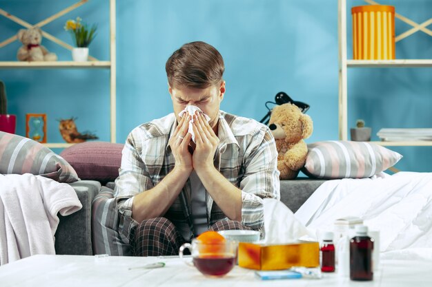 Homem barbudo doente com gripe, sentado no sofá em casa e assoando o nariz. O inverno, doença, gripe, conceito de dor. Relaxamento em casa. Conceitos de saúde.