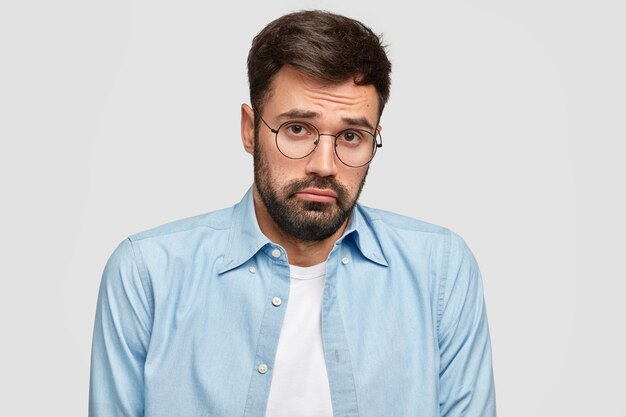 Homem barbudo e insultado confuso olha com expressão de dúvida miserável, tem que tomar decisões sérias na vida, usa óculos redondos e camisa azul, fica encostado na parede branca. Conceito de pessoas e emoções