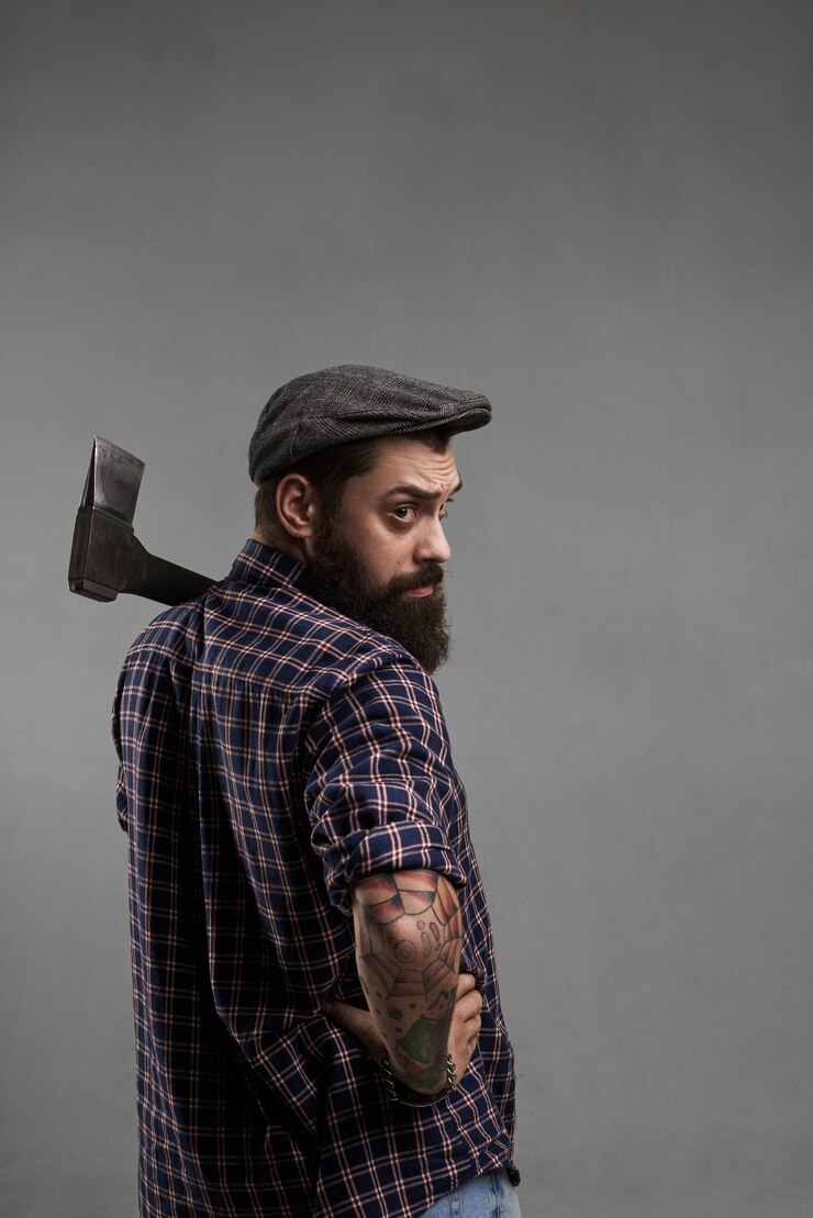 Foto grátis homem barbudo elegante com boné e camisa com machado no ombro, olha para a câmera. bonito guarda florestal filmado em estúdio. homem brutal com tatuagem.