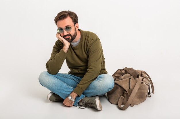 Homem barbudo elegante e bonito sentado no chão isolado vestido de moletom com bolsa de viagem, vestindo jeans e óculos escuros, triste e cansado esperando