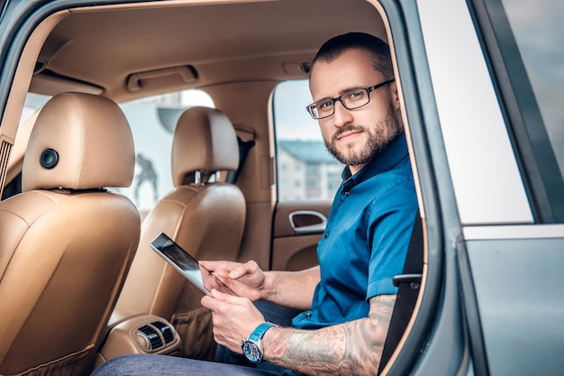 Homem barbudo elegante em óculos com tatuagem no braço usando tablet PC portátil no banco traseiro de um carro.