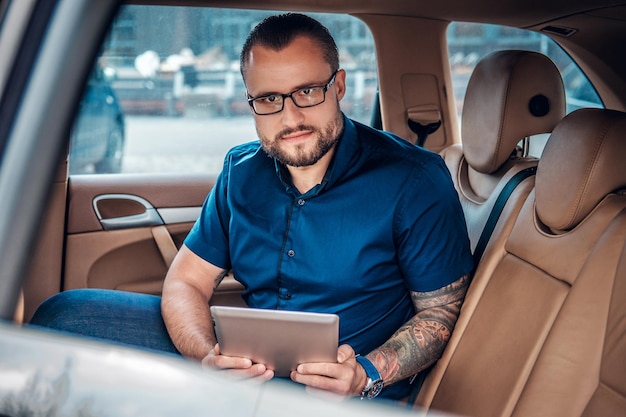 Homem barbudo elegante em óculos com tatuagem no braço usando tablet PC portátil no banco traseiro de um carro.