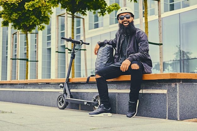 Homem barbudo elegante em óculos de sol relaxando em um banco depois de andar de scooter elétrico no centro da cidade.