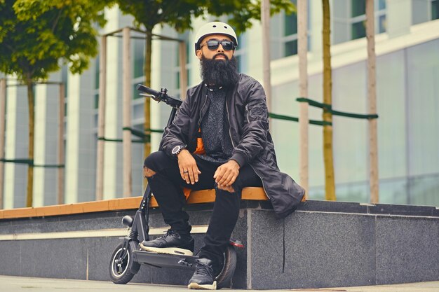 Homem barbudo elegante em óculos de sol relaxando em um banco depois de andar de scooter elétrico no centro da cidade.