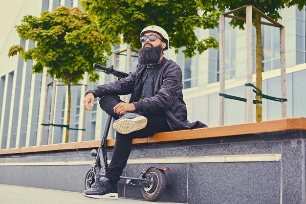 Homem barbudo elegante em óculos de sol relaxando em um banco depois de andar de scooter elétrico no centro da cidade.