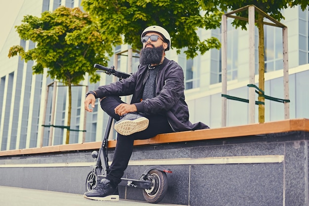 Homem barbudo elegante em óculos de sol relaxando em um banco depois de andar de scooter elétrico no centro da cidade.