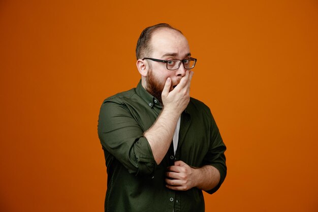 Homem barbudo em roupas casuais usando óculos olhando para a câmera espantado e chocado cobrindo a boca com a mão em pé sobre fundo laranja