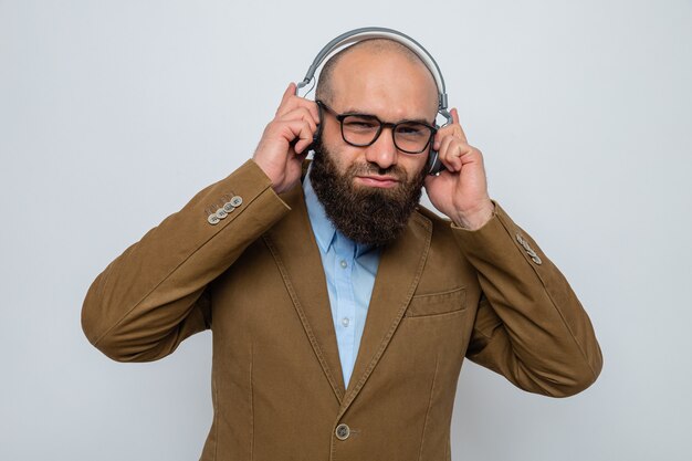 Homem barbudo em terno marrom usando óculos e fones de ouvido, sorrindo curtindo música