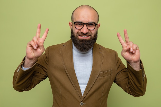 Foto grátis homem barbudo em terno marrom usando óculos, olhando para a câmera, feliz e alegre, sorrindo, mostrando o sinal v em pé sobre o fundo verde