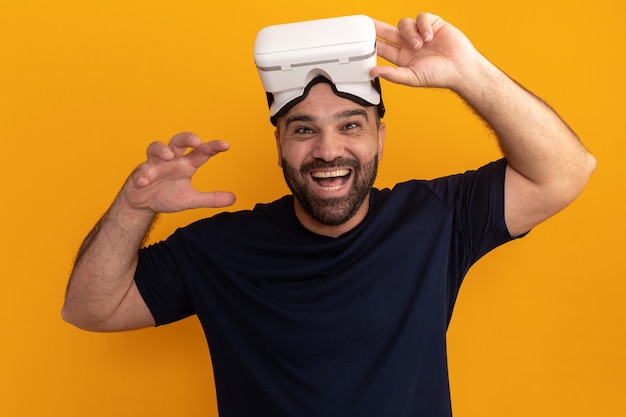 Homem barbudo em uma camiseta azul marinho com óculos de realidade virtual sorrindo alegremente com o braço levantado em pé sobre uma parede laranja